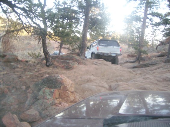 White SUV climbing rocky terrain, another vehicle visible behind trees
