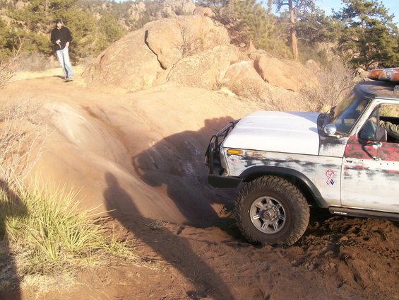 White and red 1978 Ford Bronco off-roading on a dirt trail with large rocks in the background