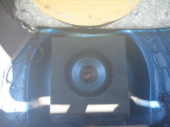 Car trunk interior with a Kenwood subwoofer in a grey box. Blue painted metal walls. Yellowish circular panel at the top