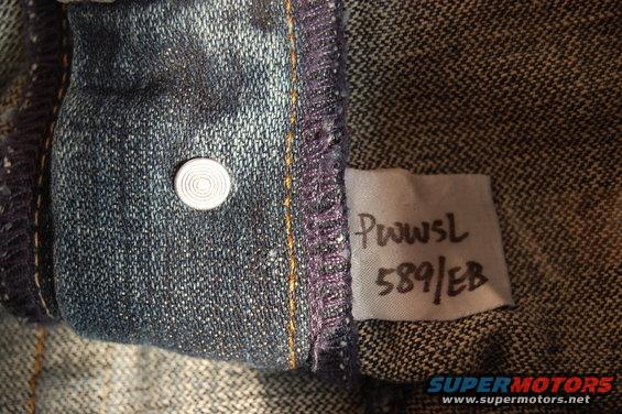 Close-up of denim fabric showing a button and a white tag with handwritten text 'PwwSL 589/EB'