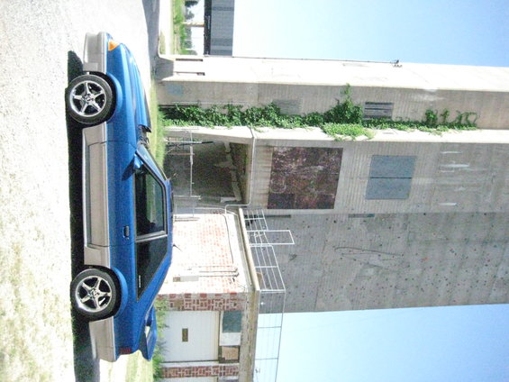 Blue two-door car with silver trim, parked on asphalt next to a brick building and a tall concrete structure