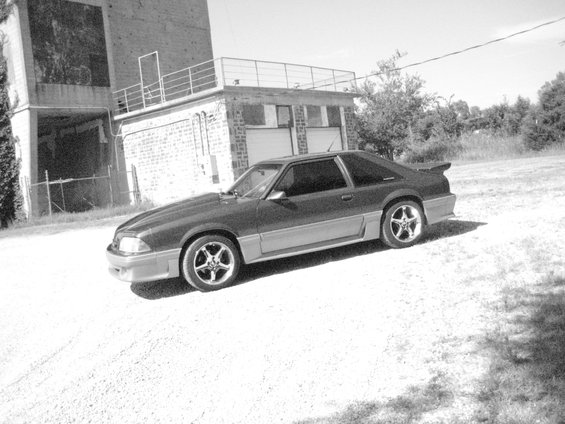 1990 Ford Mustang coupe parked on gravel in front of a brick building
