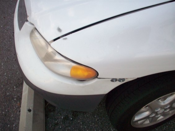 Close-up of a white car's front corner, showing the headlight and part of the bumper. The paint has some minor scuffs