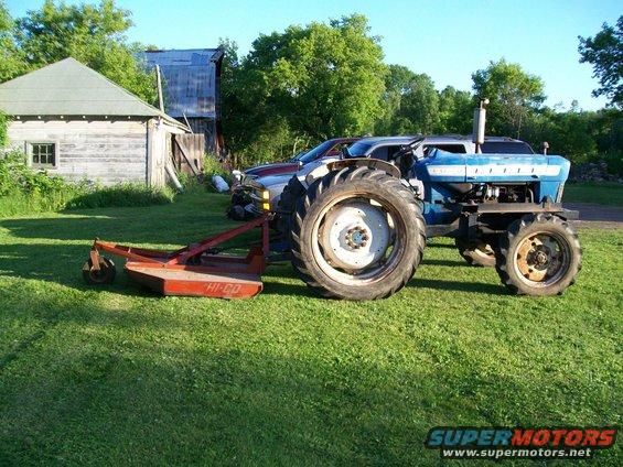 Blue tractor with a red HI-CO mower attachment parked on grass next to a building. A car is partially visible behind the tractor