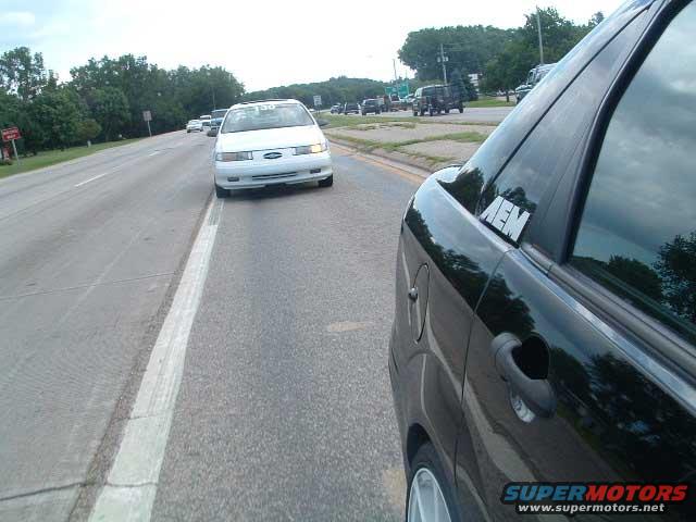 black car's side with an AEM sticker on the rear window, driving on a road next to a white car