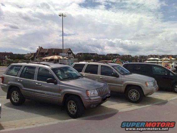 Two Jeep Grand Cherokees parked side-by-side, with a third dark vehicle behind them. Buildings and a cloudy sky are in the background