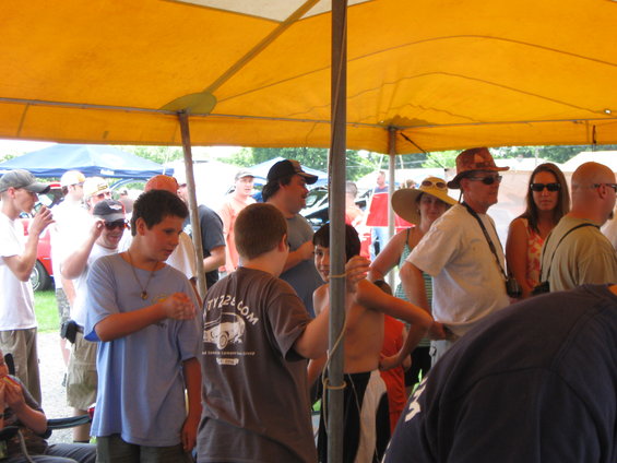 People gathered under a yellow tent at an outdoor event, with tents and trees in the background