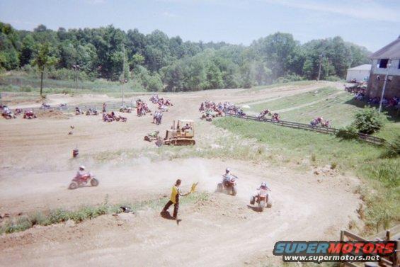 Off-road race track with ATVs, a bulldozer, and spectators. A person in yellow waves flags