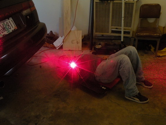 1998 Subaru Legacy rear bumper with license plate, person on creeper under car with red laser light