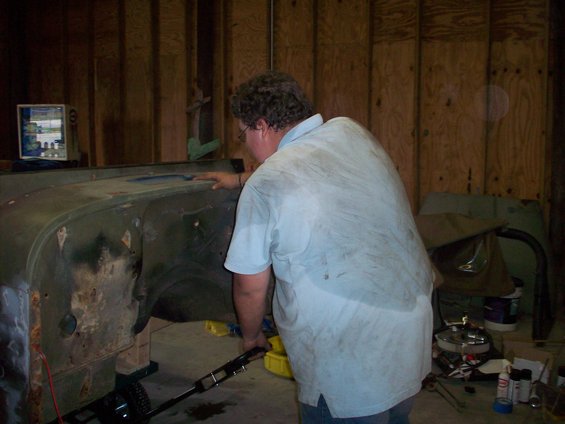 Person in light blue shirt working on the fender and inner wheel well of a green vehicle body in a garage