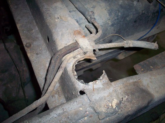 Close-up of a heavily rusted vehicle frame with fuel lines and a hole in the metal