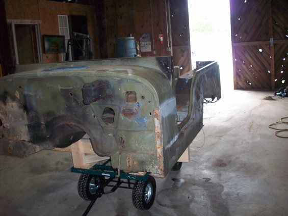 Green Jeep CJ5 body, partially disassembled and showing rust, on a green dolly inside a barn