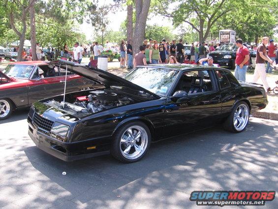 Black coupe with hood open, chrome wheels, parked at an outdoor car show. Another car is visible to the left