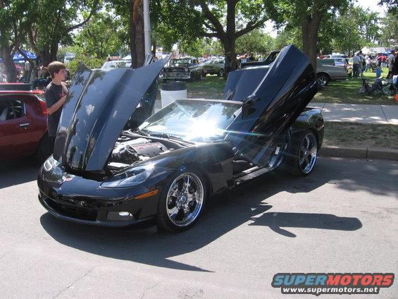Black convertible sports car with butterfly doors, hood open, chrome wheels, parked on asphalt