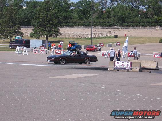 Black coupe with orange stripe on track, other cars and people visible