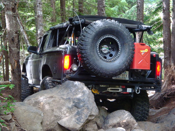 Black off-road SUV with large spare tire and red gas can on rear bumper, navigating rocky terrain