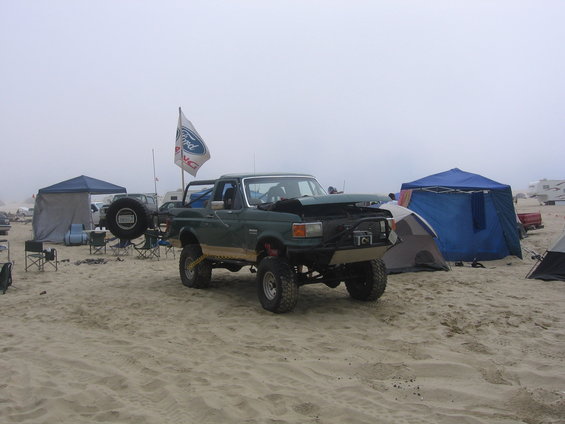 Green 1989 Ford Bronco parked on sand with aftermarket bumper, spare tire carrier, and Ford flag