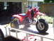 1991 Ford Bronco - A red three-wheeled ATV is secured with yellow straps to a trailer in front of a garage.
