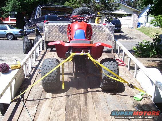 Red ATV strapped to a trailer, towed by a black pickup truck. Another car is parked nearby