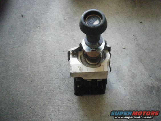 Car part with a black knob on top and a dial visible underneath. It has a metal bracket and a coiled spring