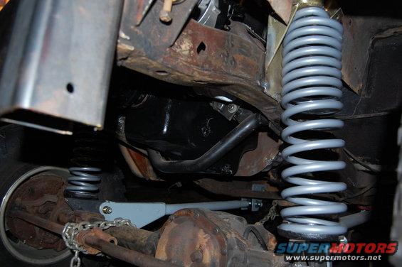 Underside view of a vehicle's suspension system, showing springs, shocks, and differential. Some rust is visible on the frame