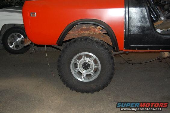 Orange and black SUV side view, showing large tire and fender. Another white vehicle is partially visible