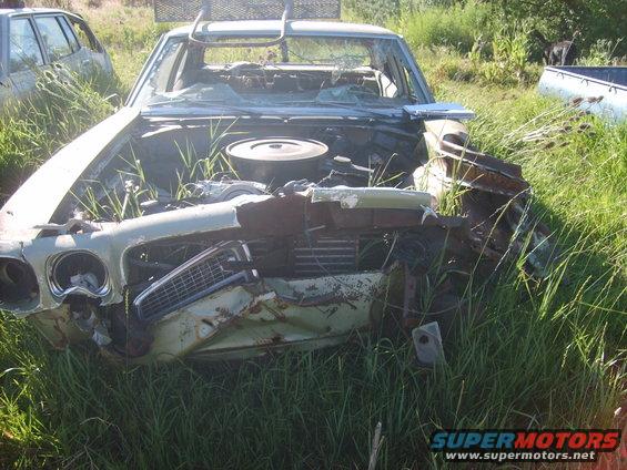 Front end of a wrecked 1970 Pontiac T-37 with its hood open, sitting in tall grass