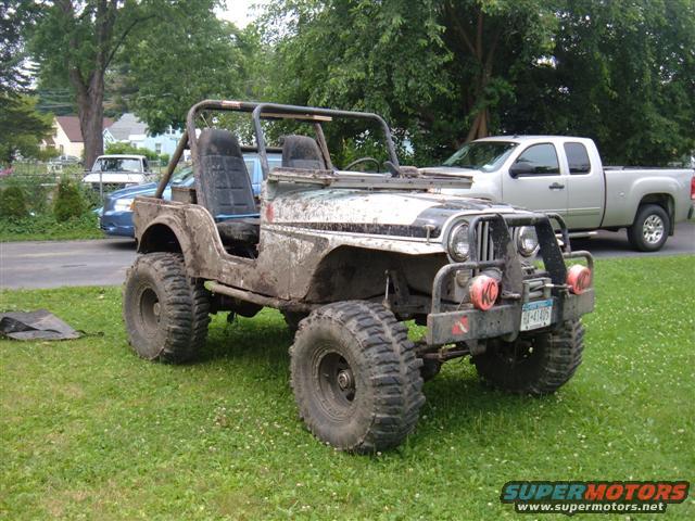 muddy Jeep with big tires parked on grass, silver pickup truck behind it