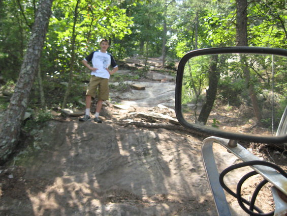 Off-road trail with a boy standing on rocks, viewed from a vehicle's side mirror
