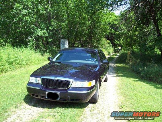 Dark purple sedan parked on a dirt road with trees and grass on either side. Front grille and headlights visible