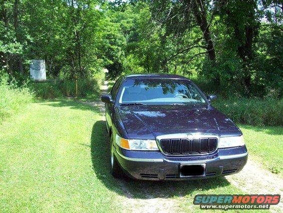 Dark sedan parked on a grassy area with trees in the background. It looks like a 1999 Mercury Grand Marquis