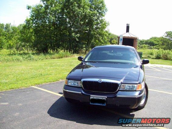 Dark sedan parked in a lot, front view. Chrome grille and trim. Trees and grass in background