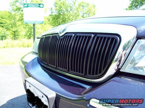 Close-up of a dark blue car's front grille with chrome trim. A sign is visible in the background