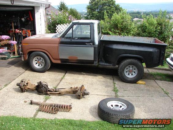 1980s Ford F-150 pickup truck parked outside a garage, with front suspension and a spare tire laid out on the ground