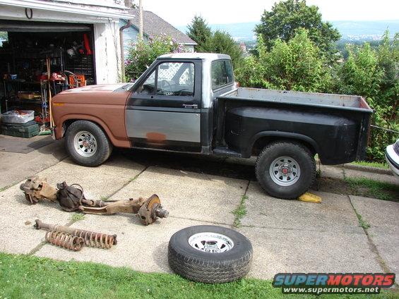 1980s Ford F-150 pickup truck, brown and silver, with front suspension parts and a spare tire on the ground in front of a garage
