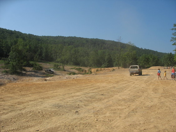 Two trucks on a dirt road with trees and hills in background. People are standing near one truck