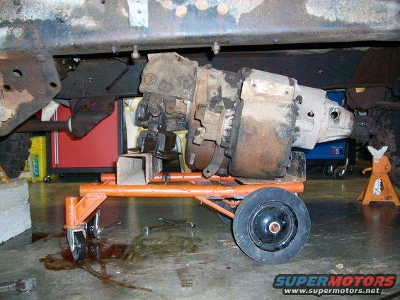 Transmission and transfer case assembly on an orange dolly, with a vehicle frame above. Floor has oil stains