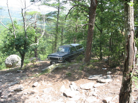 Black 1978 Ford Bronco parked on a rocky, wooded trail with a scenic overlook in the background