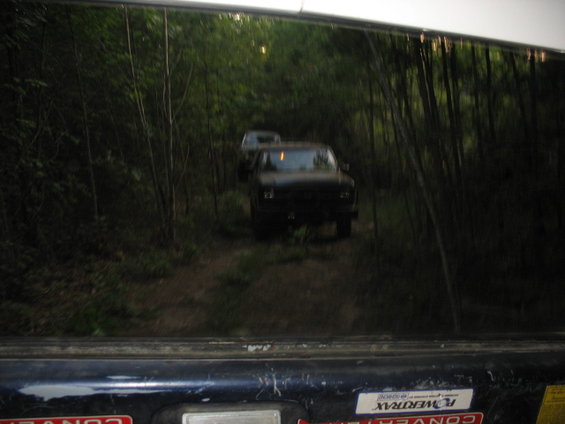 Two dark-colored trucks drive down a narrow dirt road surrounded by trees