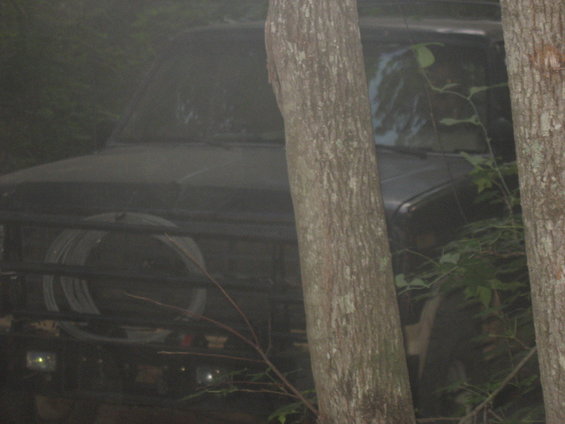 Black SUV with spare tire on front grille guard, partially obscured by trees