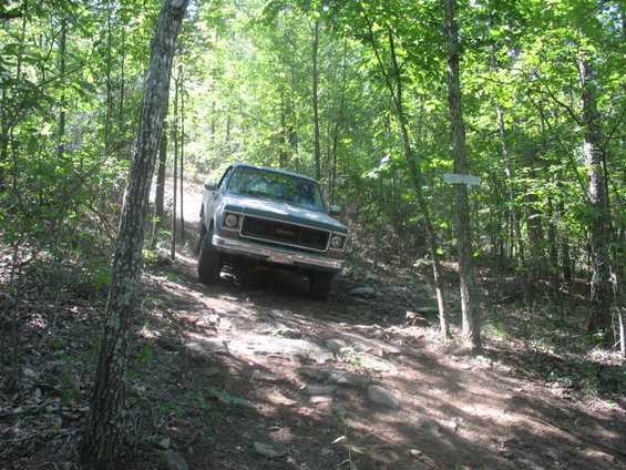GMC pickup truck driving on a rocky, wooded trail. Grille indicates mid-1970s model