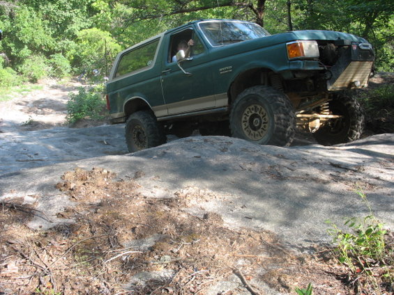 Green Ford Bronco with off-road tires climbing over rocks. Front grille is removed, showing suspension