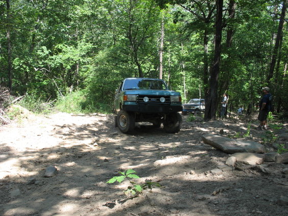 Green lifted pickup truck on a dirt path in woods, another vehicle behind it