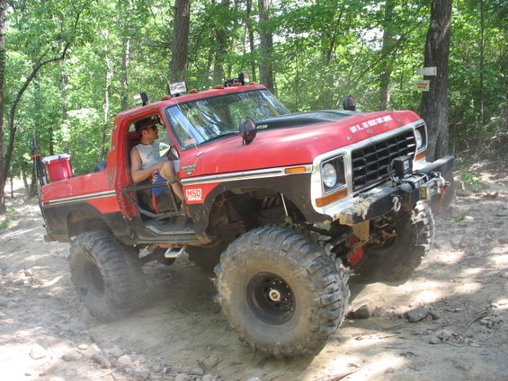 Red 1978 Ford Bronco with large off-road tires and a winch, driving on a dirt trail