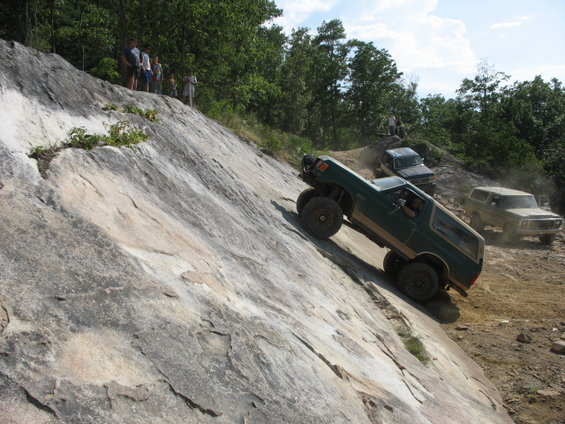 Green off-road SUV with large tires climbing a steep rock face, other vehicles and people visible on the hill