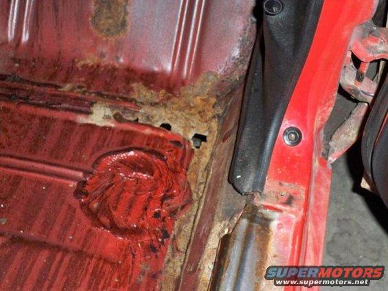 Red car interior floor pan with extensive rust and a circular patch of red material
