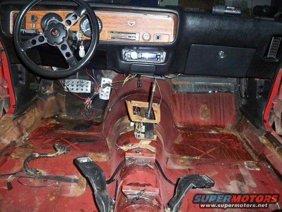 Car interior with red floor, black dashboard, and steering wheel. The floor is stripped and shows rust