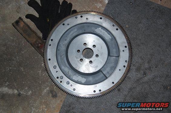 Flywheel with gear teeth around the edge, resting on a concrete floor next to black gloves and a metal bar