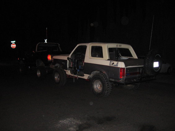 Two modified SUVs, one a two-tone Bronco, parked on a dirt road at night. The Bronco has its doors removed