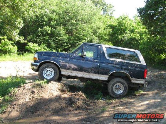 Dark blue Ford Bronco on dirt mound, trees in background